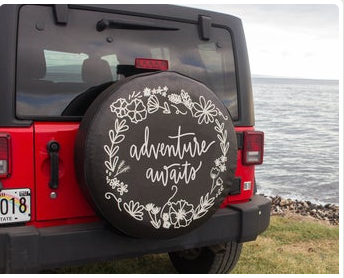 A black floral wreath tire cover with the text 'Adventure Awaits' fitted on a Jeep, with a background suggesting a beach or ocean setting.